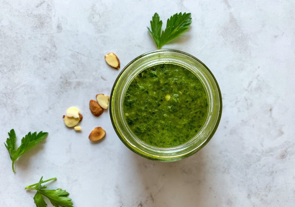 This image shows a jar of carrot top chimichurri sauce with parsley and almonds sprinkled on the ground around it. 